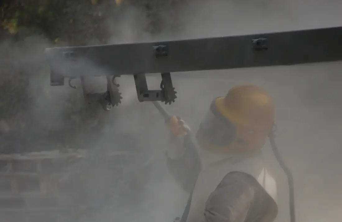 Worker in a hooded blast suit aiming a blast nozzle at a steel truck frame rail inside a cloud of dust.
