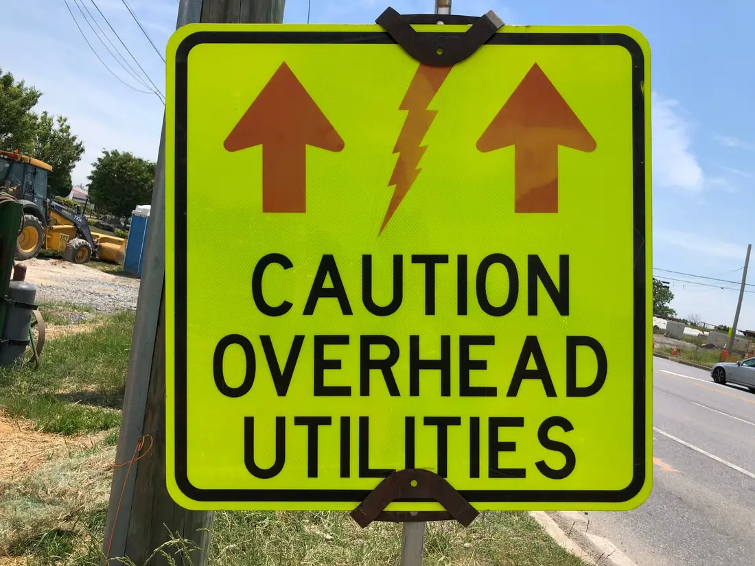 Yellow Caution Overhead Utilities sign on a pole beside a road work site, with power lines and a loader behind it.