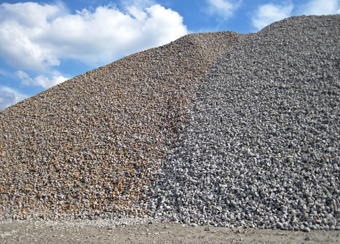 Two large quarry stockpiles of angular crushed limestone, one tan and one gray, under a blue sky.