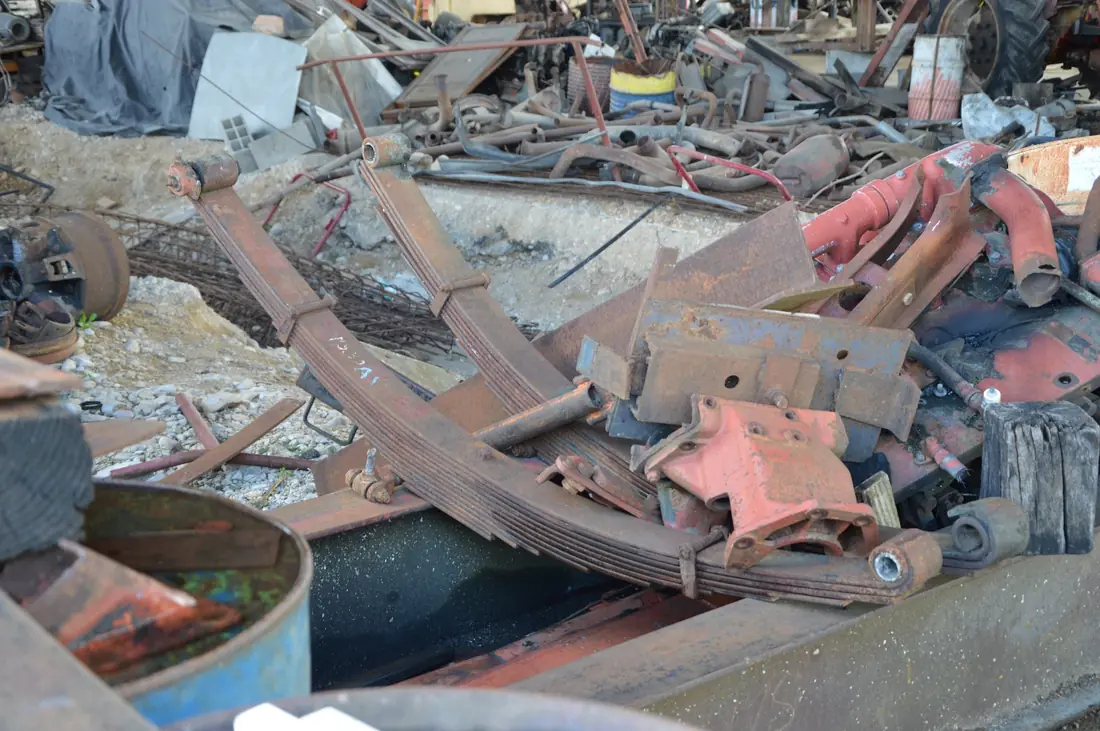 Two rusty multi-leaf springs, each with a rolled eye at both ends, lying on scrap metal in a salvage yard.