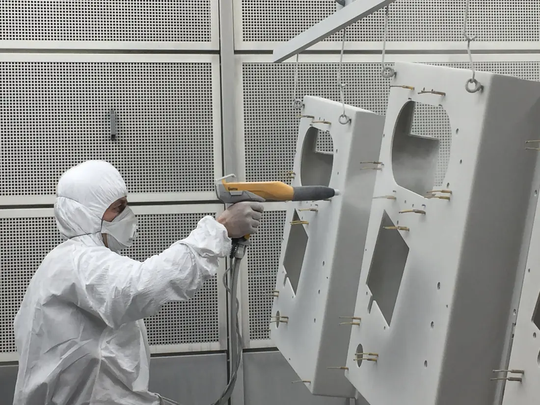 Worker in a white suit and mask spraying powder from a coating gun onto steel parts hanging from hooks in a booth.