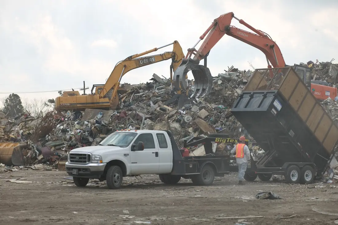 Pickup with a gooseneck dump trailer raised to unload debris at a disposal site while excavators work the pile behind it.
