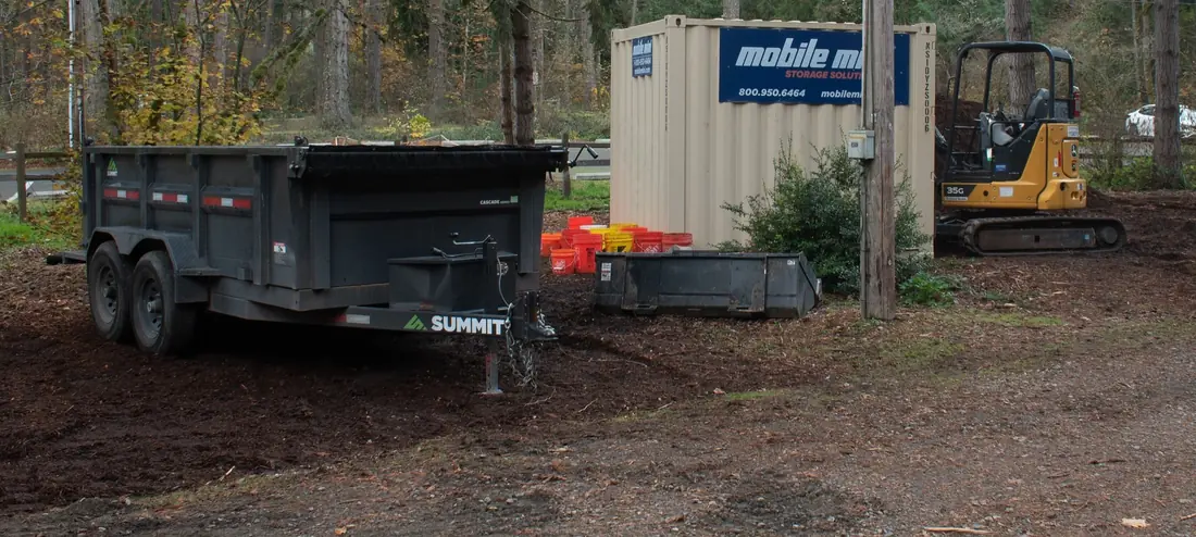 Tandem-axle bumper-pull dump trailer parked on its jack at a wooded job site, with a mini excavator nearby.