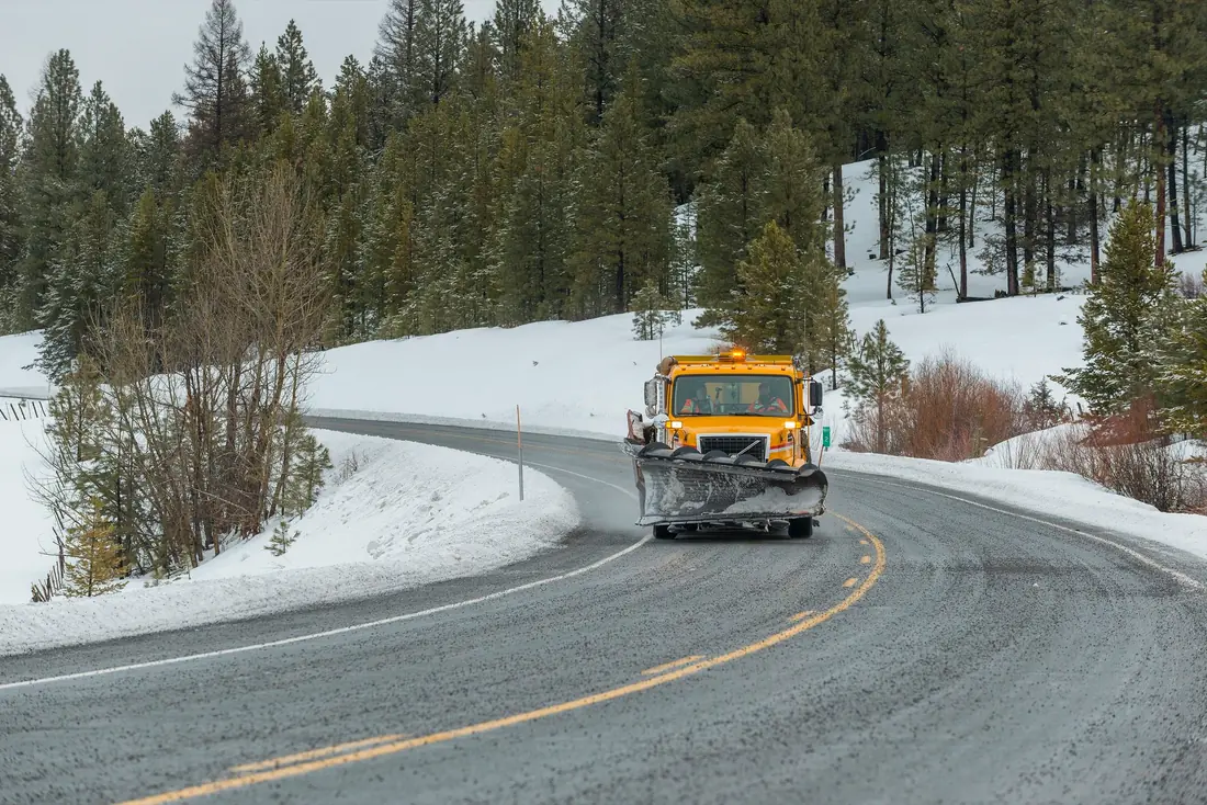 Yellow plow truck driving a two-lane highway between snowbanks, the pavement grey with salt and road film.