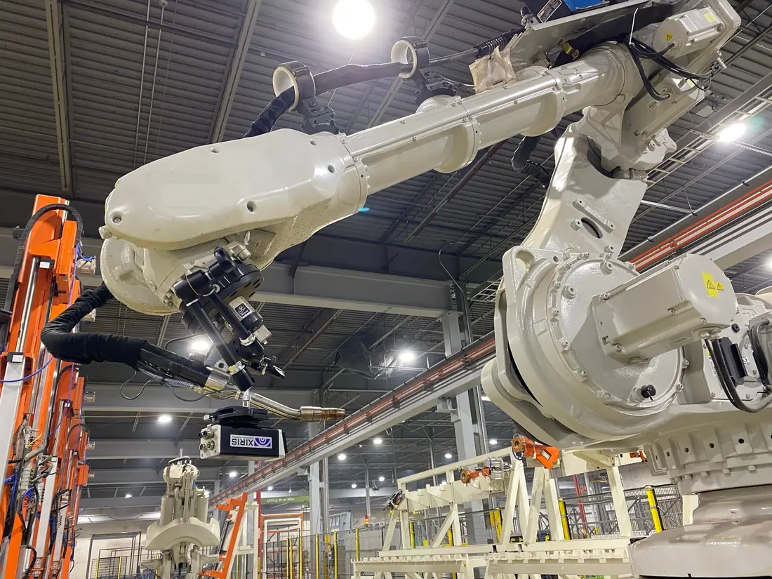 Industrial robot arm holding a welding torch over a work fixture in a factory, with a weld camera mounted beside the torch.