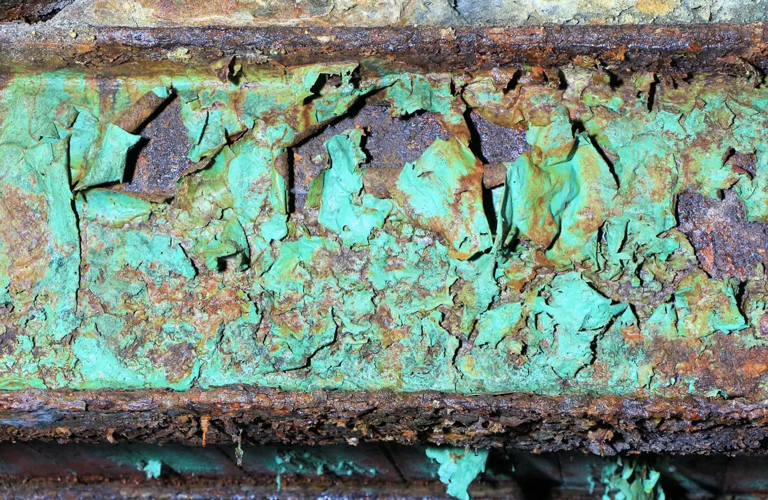Close-up of a steel beam where rust has lifted green paint off in curled sheets, exposing scaly brown steel.