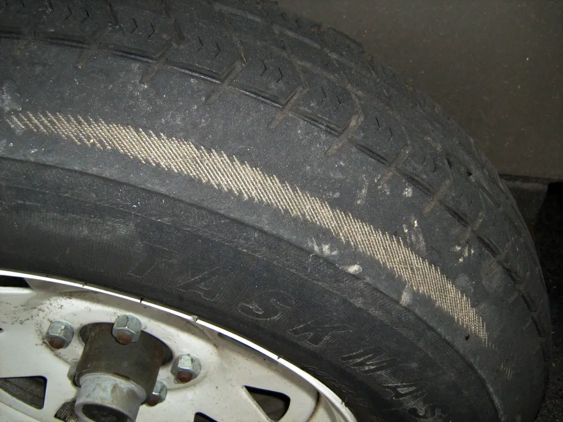 Tire on a white trailer wheel with a band of fabric cords showing where the rubber has worn through near the shoulder.